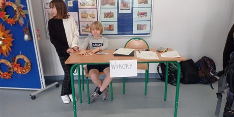 Powiększ grafikę: Dwie osoby na korytarzu szkolnym są w komisji. Po lewej stronie osoba stoi a po prawej siedzi.